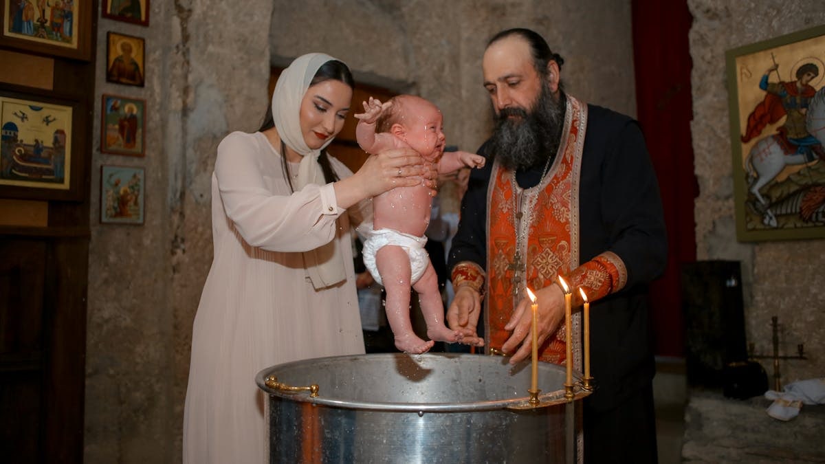A baby being baptized by a priest and woman in a traditional church setting.