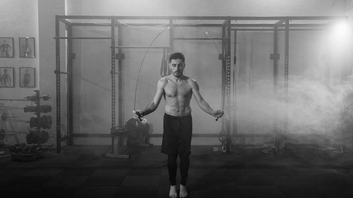 Muscular man doing a jump rope workout in an industrial gym setting with dramatic smoke around.