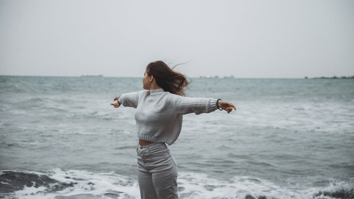 A woman stands with arms spread by the sea, embracing the freedom of a windy, overcast day.
